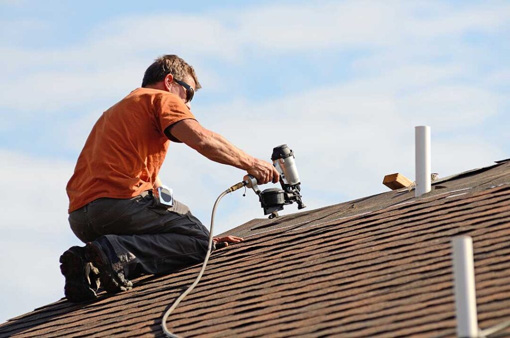 Homeowner inspecting roof shingles and gutters beside a snow-dusted house with a ladder and fallen leaves nearby