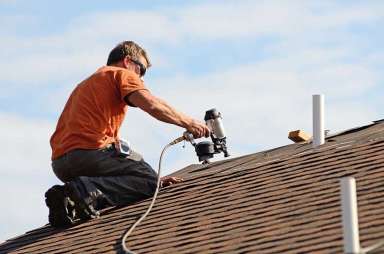 Homeowner inspecting roof shingles and gutters beside a snow-dusted house with a ladder and fallen leaves nearby